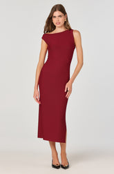 A woman is wearing a sleeveless, full-length, deep red dress with a boat neckline. The dress is fitted, accentuating her figure, and features a side slit at the hem. She is accessorized with gold hoop earrings and rings on her fingers. She is wearing black high heels with metallic accents on the toes.